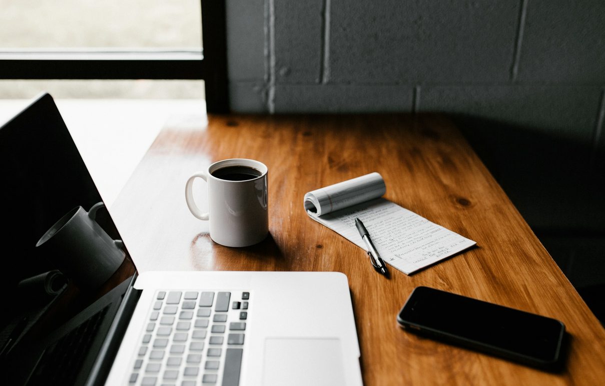 A desk with a notebook, a laptop and a cup of coffee