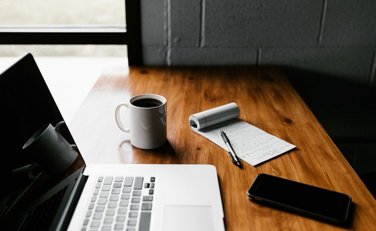A desk with a notebook, a laptop and a cup of coffee